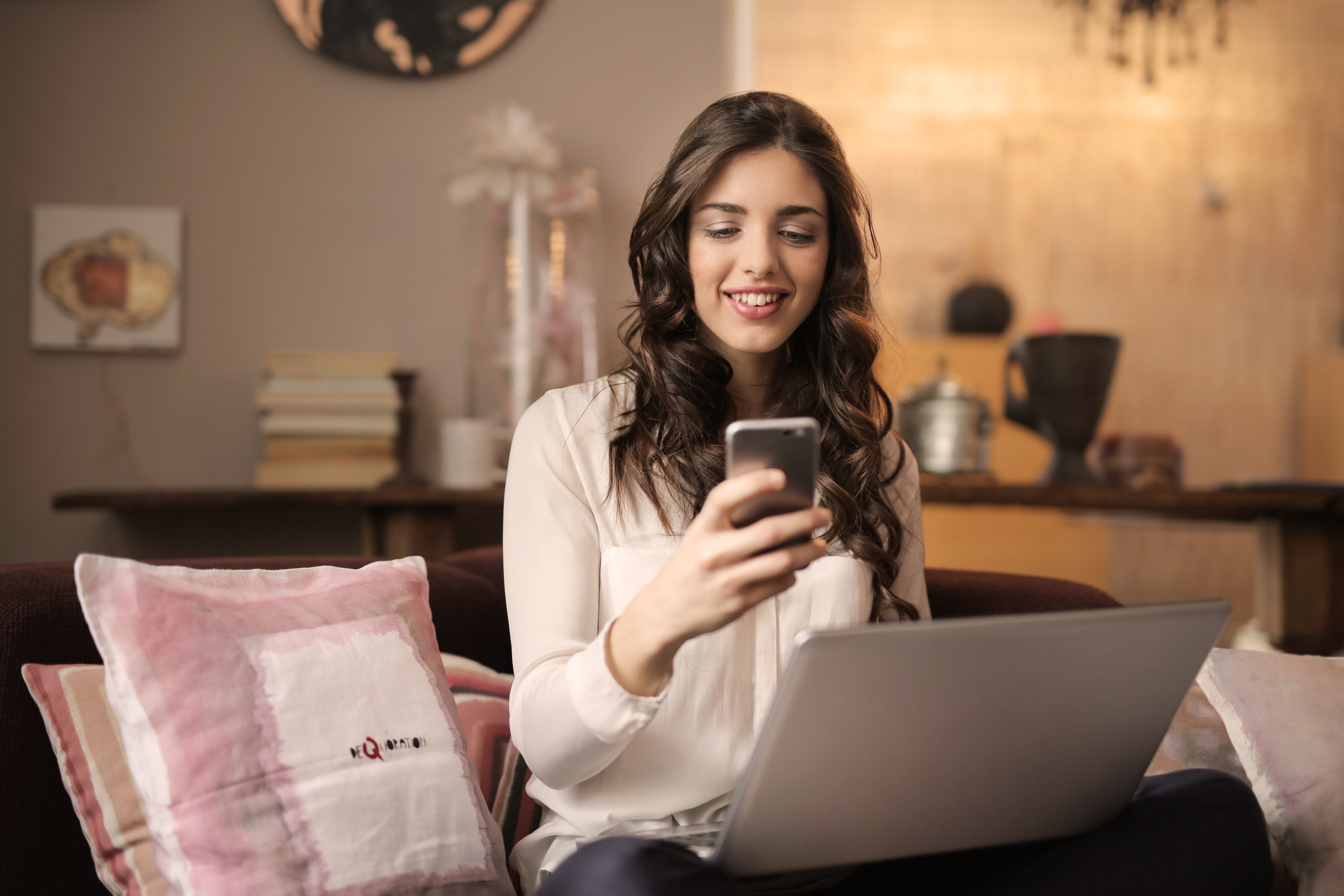 Photograph showing woman sitting on sofa with laptop on her lap and phone in her hand